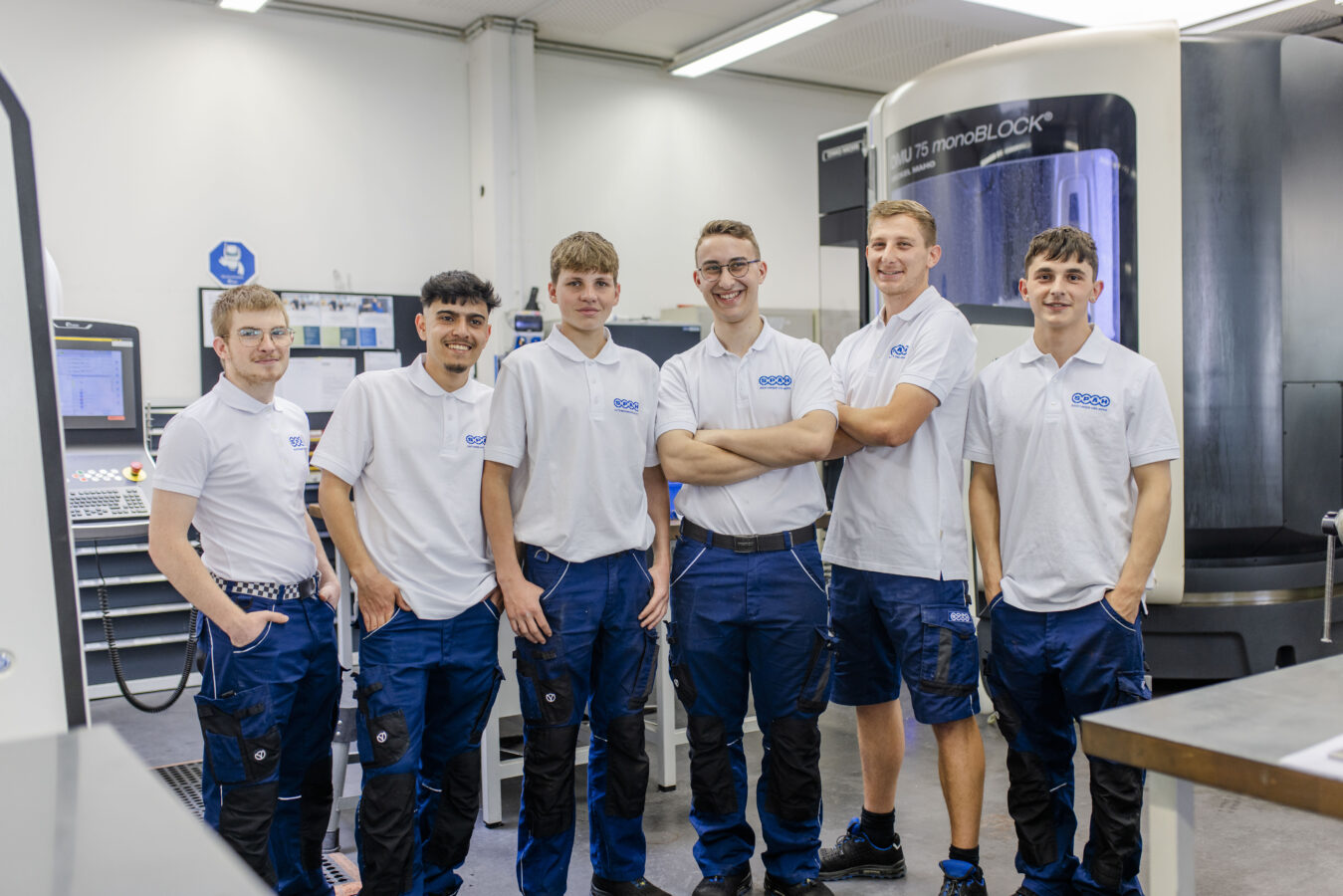 Das Bild zeigt eine Gruppe von sechs lächelnden gewerblichen Azubis in weißen Poloshirts mit Firmenlogo und blauer Arbeitshose, die selbstbewusst mit den Händen in den Taschen oder verschränkten Armen in einer hellen Werkstatt vor einer CNC-Maschine und einem Maschinenbedienfeld posieren.