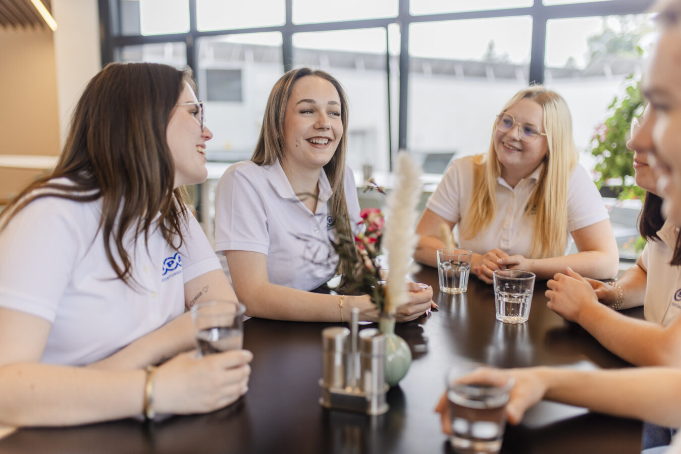 Das Bild zeigt eine Gruppe Azubis in weißen Poloshirts, die an einem schwarzen Tisch mit Wassergläsern sitzen und sich austauschen, wobei alle freundlich lächeln.