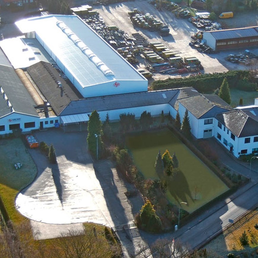 Eine Luftaufnahme zeigt das Firmengebäude der Karl Popp GmbH im Jahr 1972 mit einem langen Produktionshallen-Anbau und einem gepflegten Rasenbereich, umgeben von einem großen Lagerbereich mit Fahrzeugen und Material.