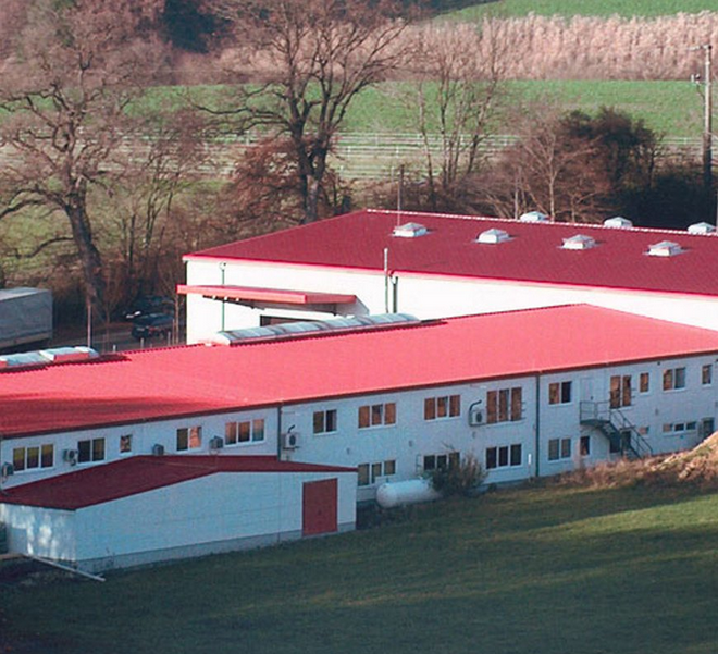 Das Bild zeigt eine von Bäumen umgebene Industriefläche mit mehreren weißen, ein- und zweistöckigen Gebäudeteilen mit auffälligen, rot gedeckten Dächern.