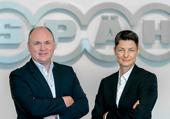 Das Business-Porträt zeigt rechts Sandra Späh und links Dr. Wolfgang Hergeth in dunklen Anzügen und mit verschränkten Armen vor dem großen, silberfarbenen Späh-Logo.