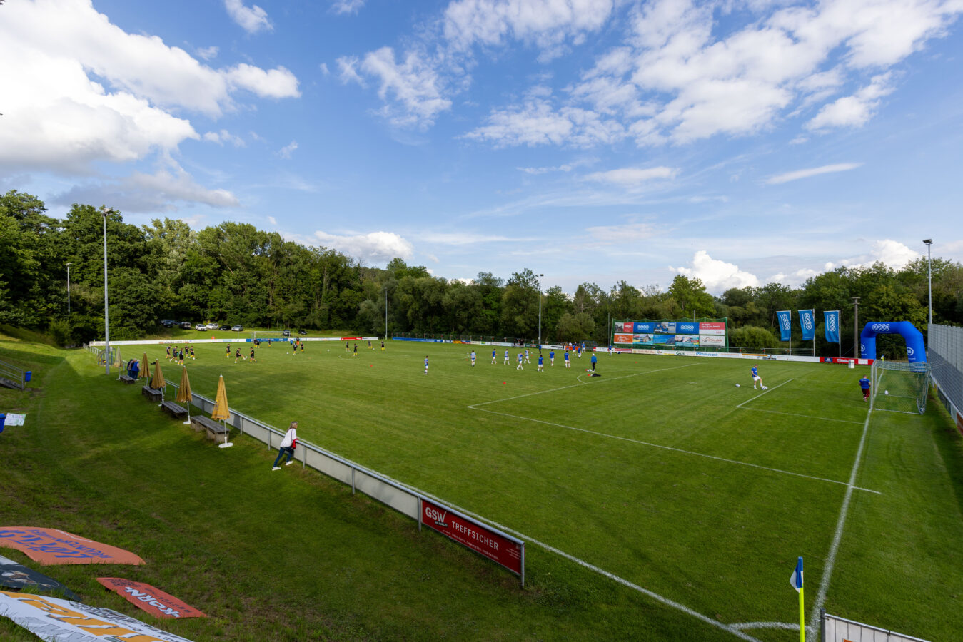 Eine Weitwinkelaufnahme zeigt den belebten Fußballplatz während des Firmenturniers unter einem leicht bewölkten Himmel.