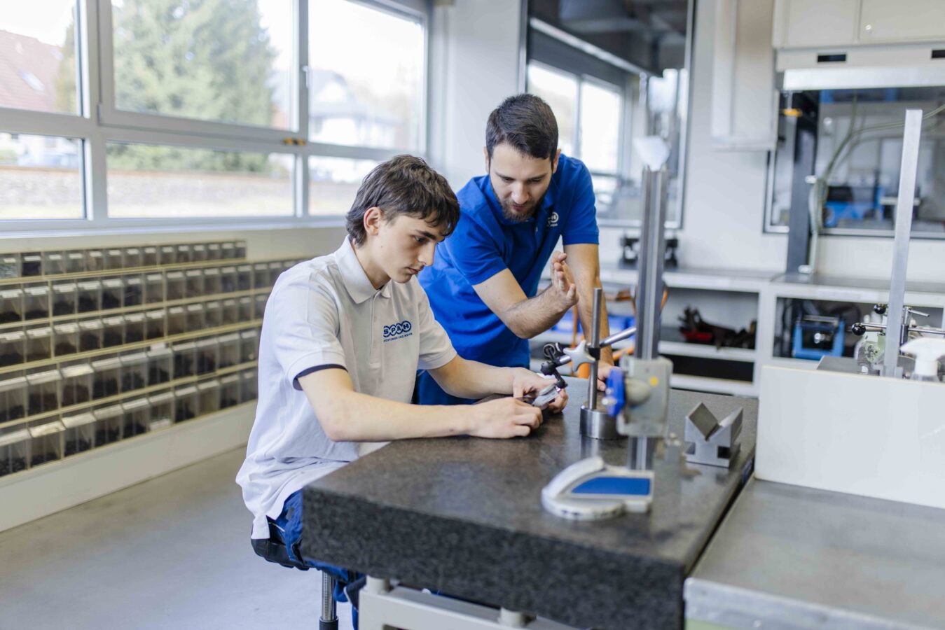 Das Bild zeigt einen Azubi, der konzentriert ein Werkstück auf einer dunklen Messplatte betrachtet, während der Ausbilder ihm aufmerksam zusieht und etwas erklärt.