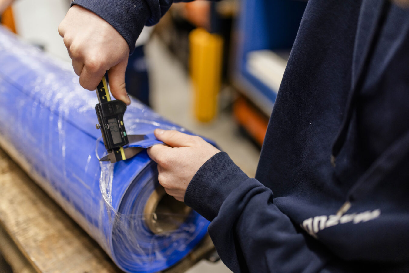Eine Nahaufnahme zeigt zwei Hände, die mit einem digitalen Messschieber die Dicke einer großen, in Plastik eingewickelten Rolle aus blauem Material messen.