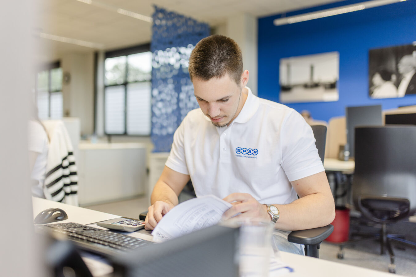 Das Bild zeigt einen jungen, konzentrierten Werkstudenten im weißen Poloshirt, der an einem hellen Schreibtisch in einem modernen Büro mit blauen Wänden sitzt und Unterlagen durchsieht.