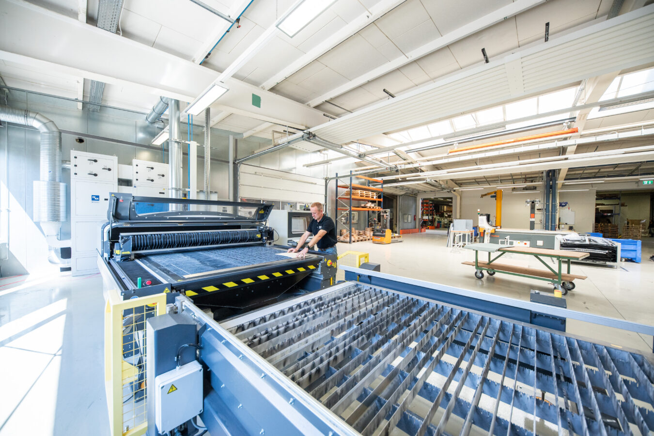 Ein Mitarbeiter bedient in einer hellen, weitläufigen Werkhalle eine große, industrielle Laserschneidanlage zur Bearbeitung von Plattenmaterial.