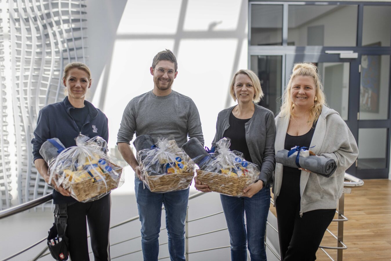 Vier Späh-Mitarbeiter stehen lächelnd in einem modernen Innenbereich mit viel Tageslicht und halten Geschenkkörbe mit Decken und anderen verpackten Gegenständen, die sie offenbar als Anerkennung für ihre Leistungen erhalten haben.