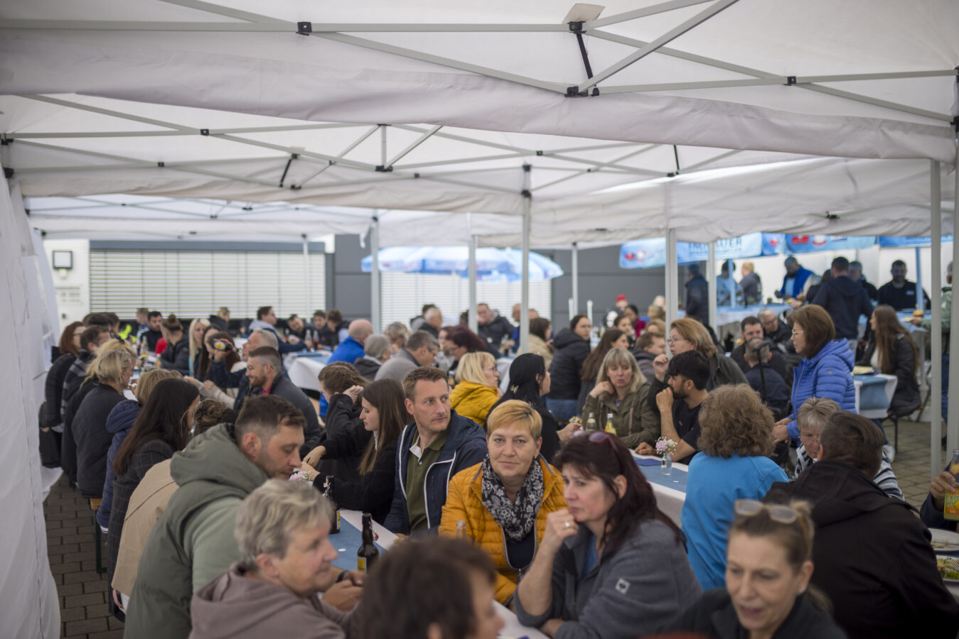 Zahlreiche Späh-Mitarbeiter und ihre Gäste sitzen dicht gedrängt an langen Tischen unter einem großen Partyzelt im Freien beim Grillfest.