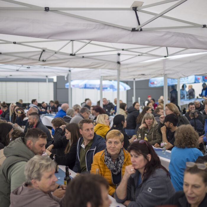 Zahlreiche Späh-Mitarbeiter und ihre Gäste sitzen dicht gedrängt an langen Tischen unter einem großen Partyzelt im Freien beim Grillfest.