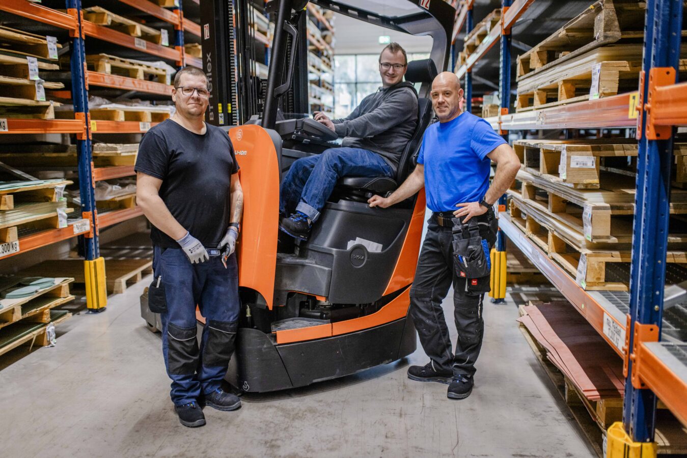 Drei lächelnde Späh-Mitarbeiter stehen in einem Lagergang mit vollen Hochregalen: Ein Mann in einem schwarzen T-Shirt und Arbeitshandschuhen steht links, ein weiterer Mann mit Brille sitzt auf einem orangefarbenen Stapler in der Mitte, und ein dritter Mann in einem blauen T-Shirt und schwarzer Arbeitshose steht rechts mit den Händen in die Hüften gestemmt.