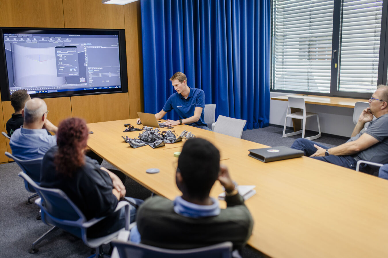In einem Besprechungsraum arbeitet ein Mitarbeiter an einem Laptop vor einem großen Bildschirm, der eine CAD-Software zur 3D-Modellierung zeigt.