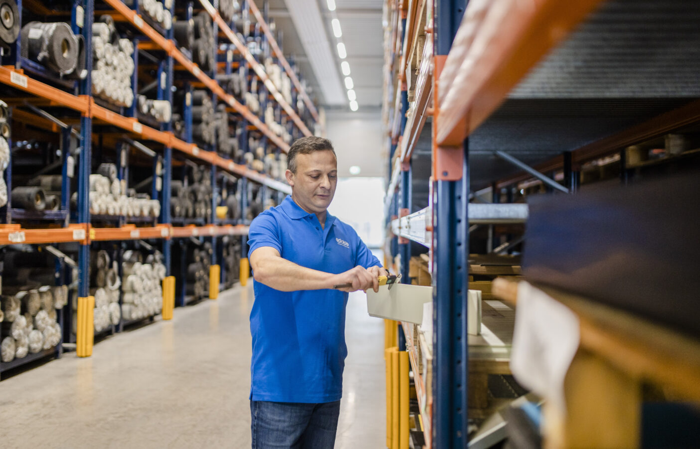 Ein Mitarbeiter von Späh in einem blauen Polohemd entnimmt in einem Lager mit Hochregalen eine Materialprobe zur Qualitätsprüfung.