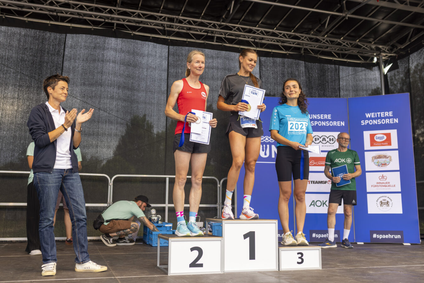 Drei Läuferinnen stehen bei einer Siegerehrung auf einem Podest und halten ihre Urkunden und Medaillen in den Händen.