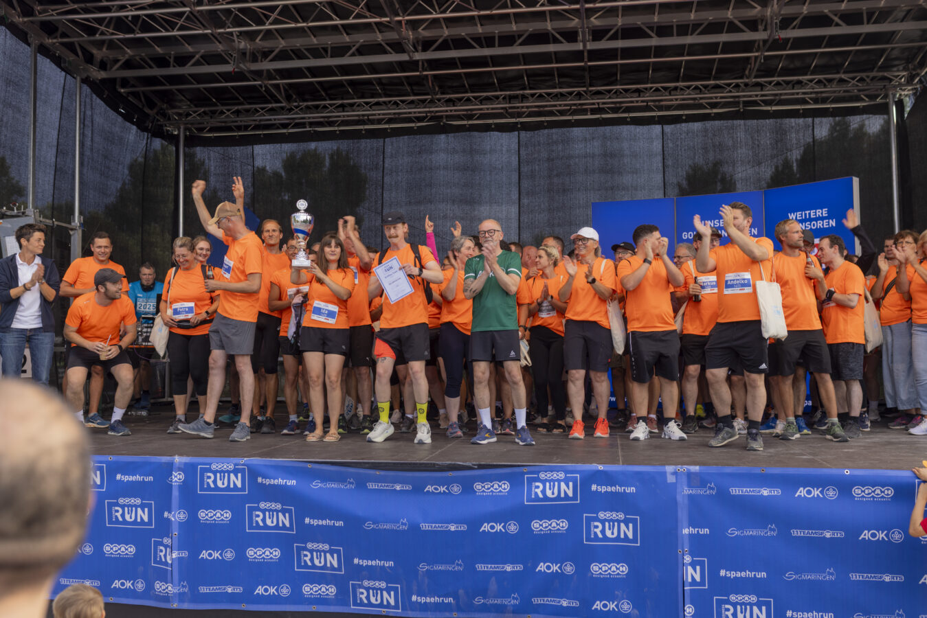 Eine große Gruppe von Läufern in orangefarbenen T-Shirts feiert gemeinsam auf einer Bühne und präsentiert stolz einen gewonnenen Pokal.