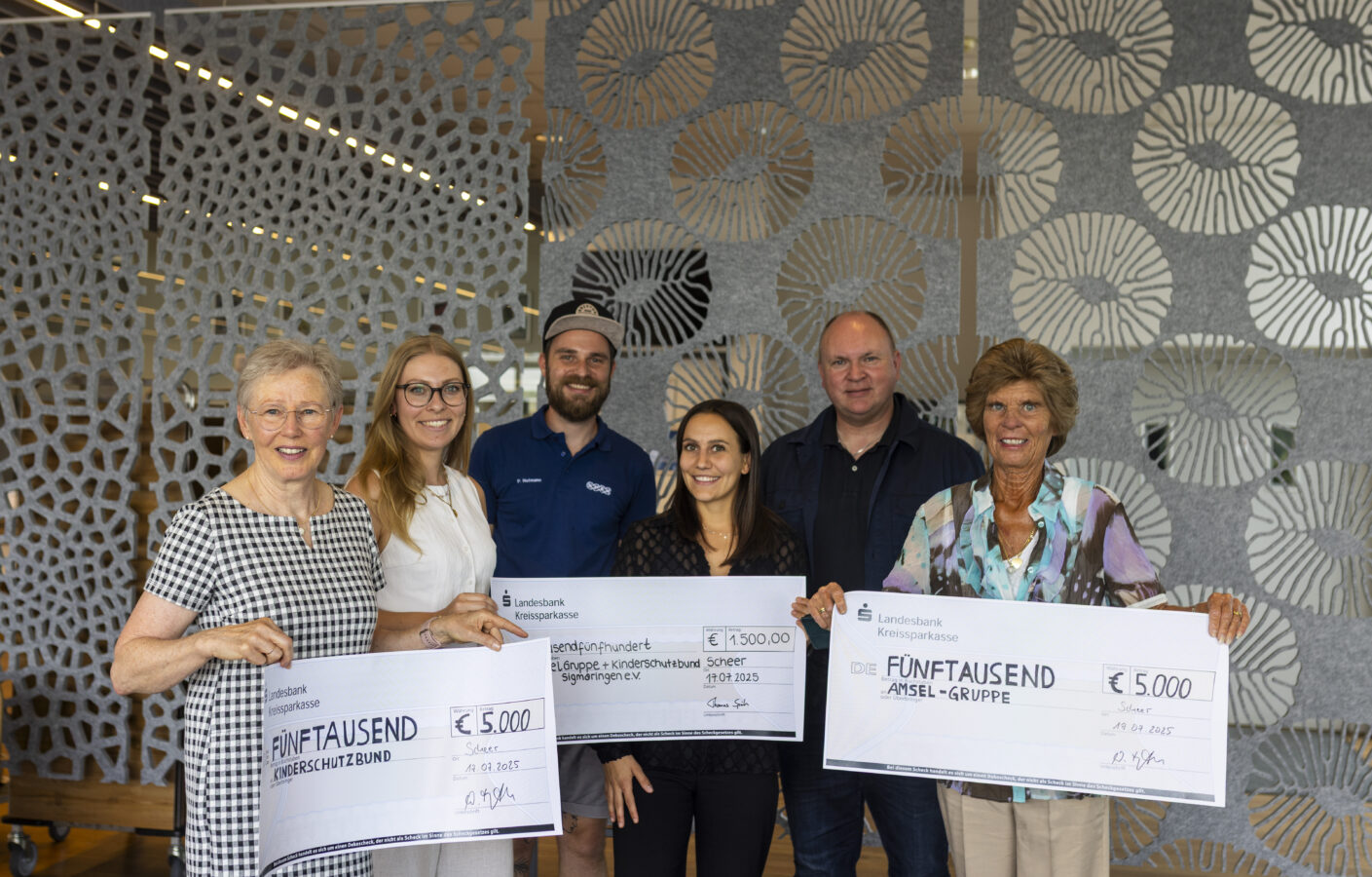 Eine Gruppe von sechs Personen, drei Frauen und drei Männer, hält Schecks der Landesbank Kreissparkasse im Wert von € 5.000 für den Kinderschutzbund und die Amsel-Gruppe sowie einen Scheck über € 1.500 für die Jugend- und Kinderkrebshilfe Sigmaringen in einem modernen Innenraum mit einer gemusterten Trennwand im Hintergrund.