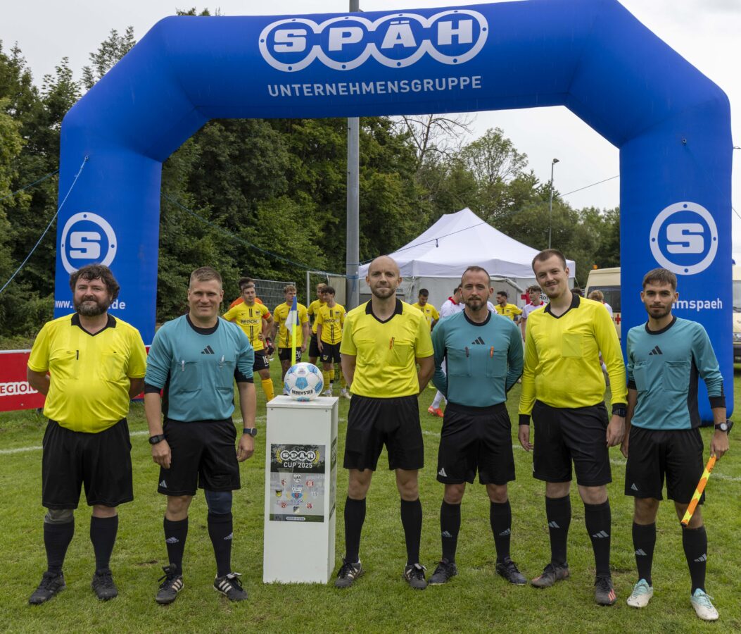 Sechs Mitspieler des Turniers posieren in gelben und türkisen Shirts vor dem Anpfiff für ein Gruppenfoto auf dem Rasen.