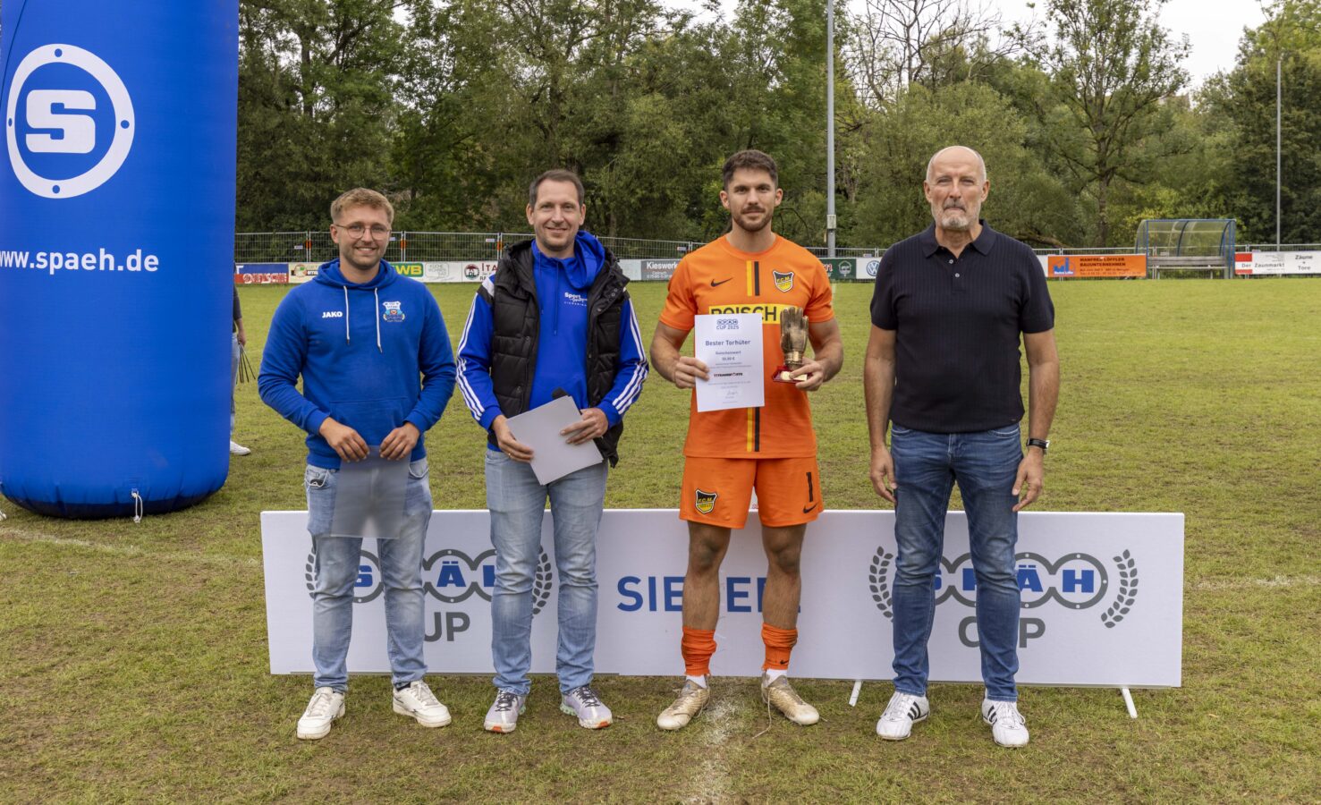 Ein Fußballspieler im orangefarbenen Trikot nimmt eine Auszeichnung als bester Torhüter entgegen, flankiert von drei Organisatoren.