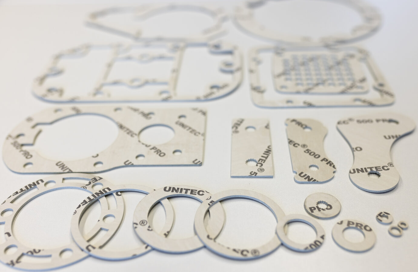 Das Bild zeigt eine Auswahl von hellbeigen UNITEC® 500 PRO Dichtungen in verschiedenen kreisförmigen und komplexeren Formen auf einer weißen Oberfläche.