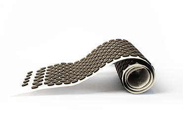 Eine weiße Trägerfolie, auf der eine Vielzahl kleiner, runder Dämpfungspads aus schwarzem Schaumstoff aufgebracht sind, ist teilweise aufgerollt.