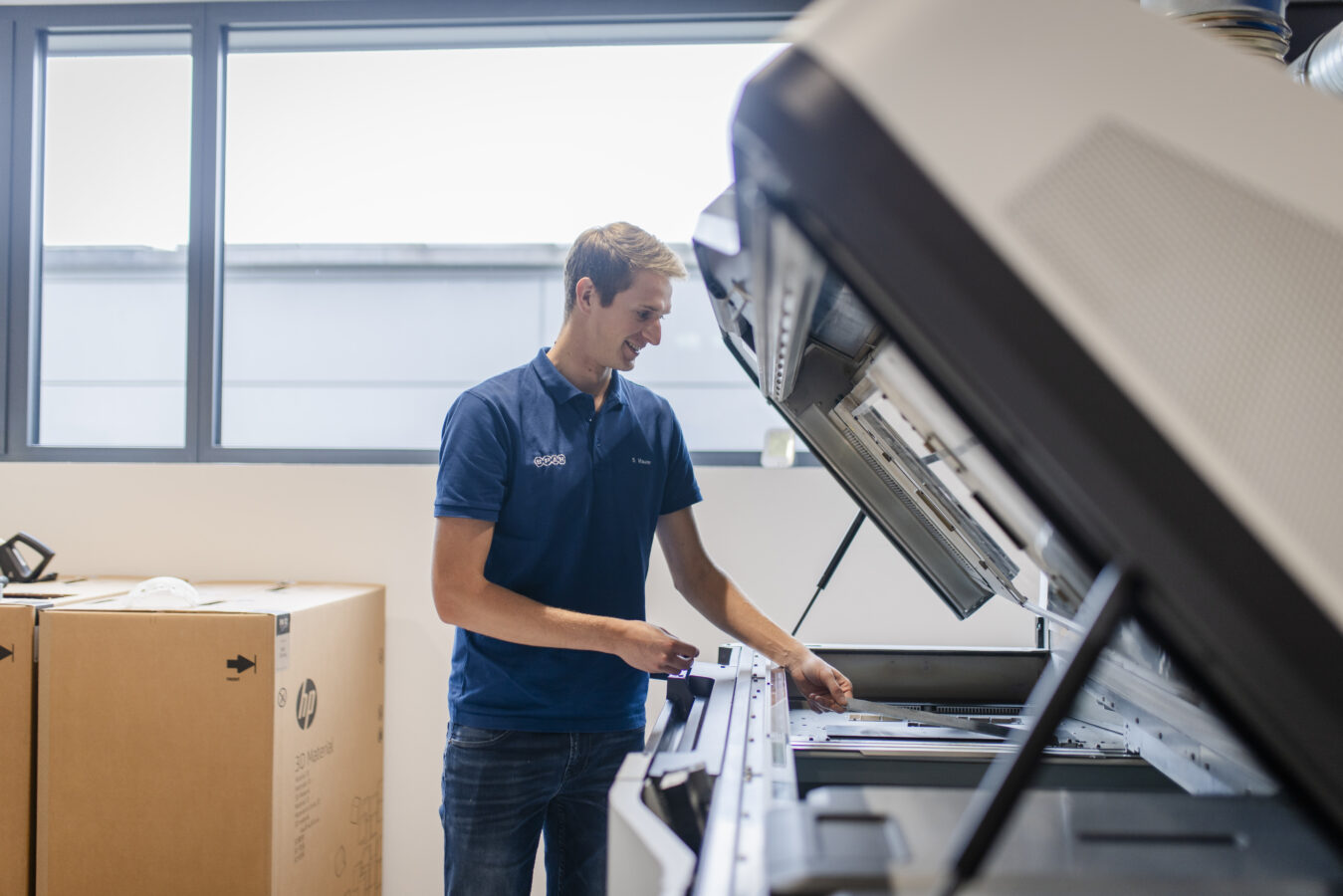 Das Bild zeigt einen lächelnden Mitarbeiter der Firma Späh in einem blauen Polohemd, der an einer geöffneten industriellen 3D-Druckanlage arbeitet.