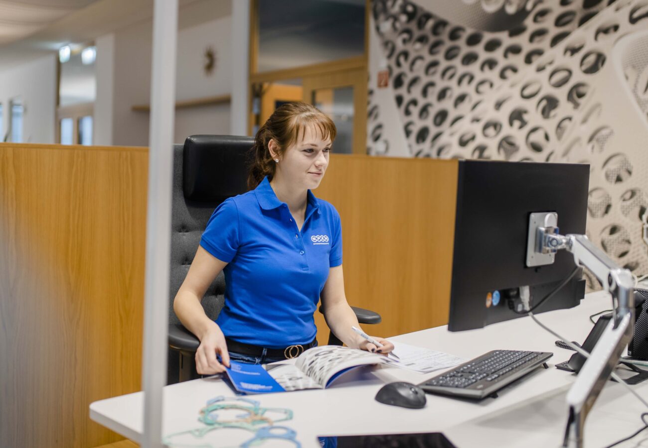 Das Bild zeigt eine Späh-Mitarbeiterin in einem blauen Poloshirt, die an einem modernen Schreibtisch vor einem Computermonitor sitzt und sich auf ihre Arbeit konzentriert, während sie einen Katalog durchsieht.