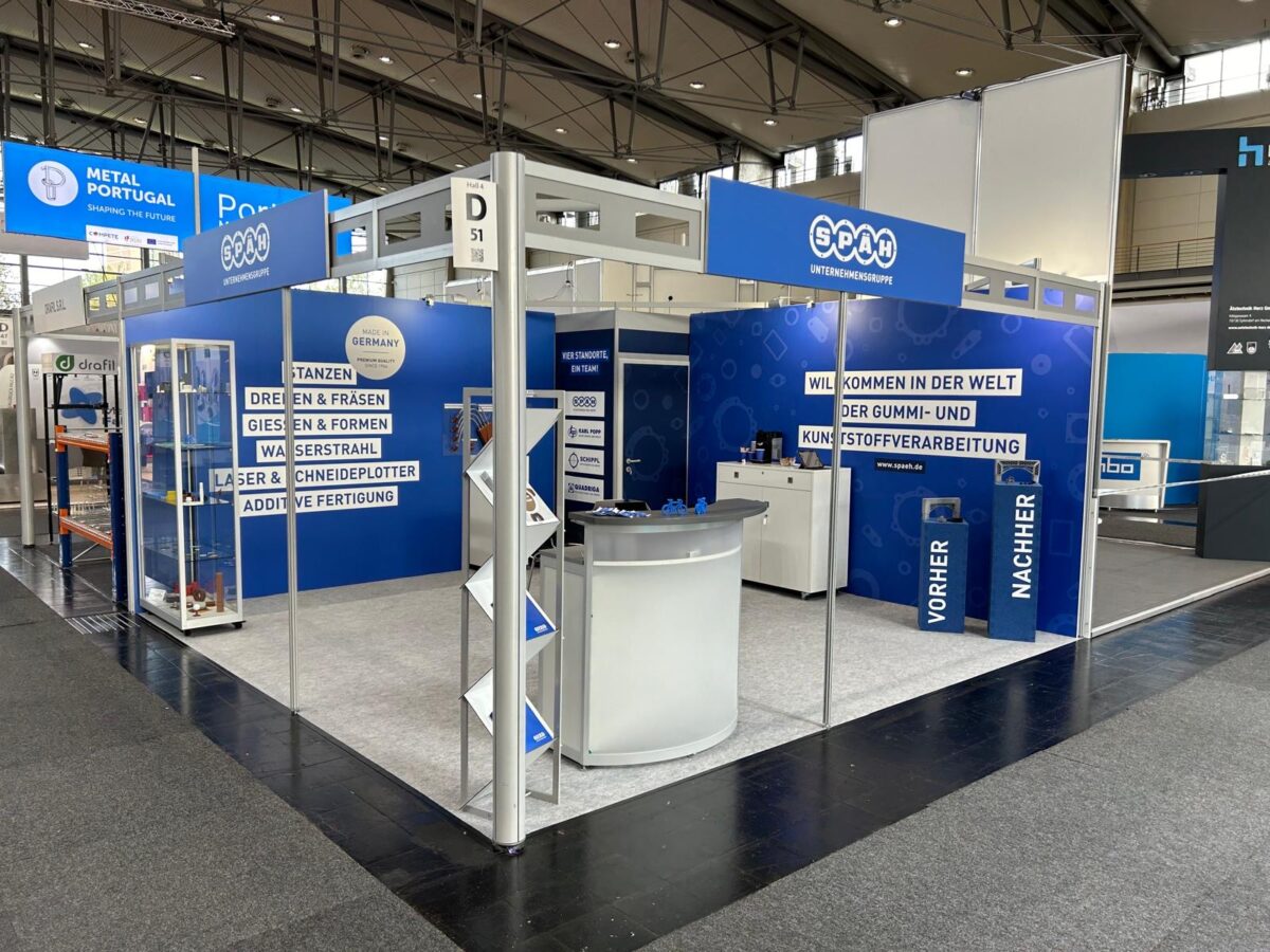 Das Bild zeigt einen großflächigen Messestand der Firma Späh in den Farben Blau und Weiß, der verschiedene Dienstleistungen im Bereich Gummi- und Kunststoffverarbeitung bewirbt und einen Empfangstresen, eine Regalwand mit Exponaten und große Banner mit Aufschriften wie "DREHEN & FRÄSEN" und "WILLKOMMEN IN DER WELT DER GUMMI- UND KUNSTSTOFFVERARBEITUNG" präsentiert.