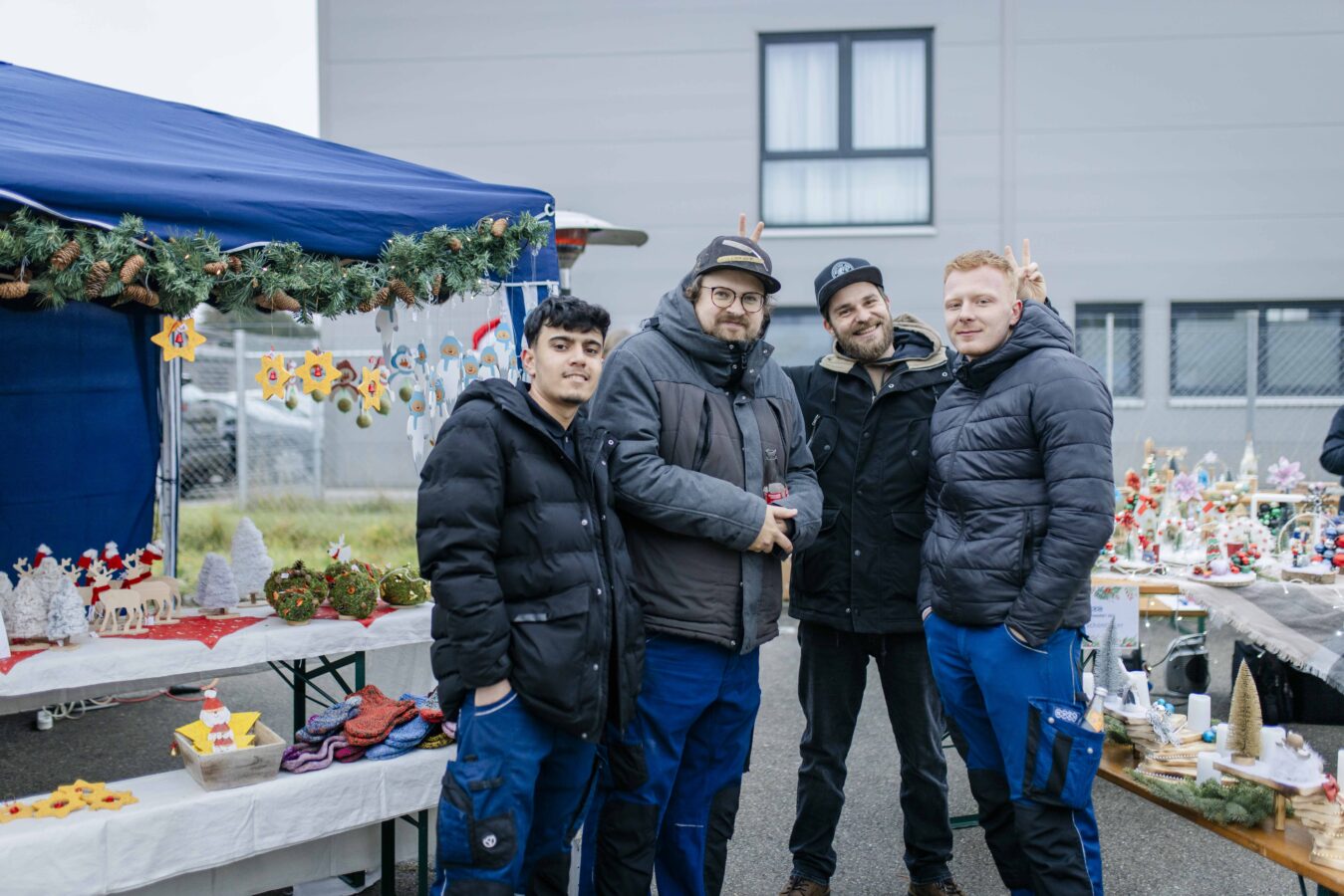 Vier Männer stehen gut gelaunt vor einem blauen Marktstand, der mit Weihnachtsdekoration und kleinen Figuren geschmückt ist.