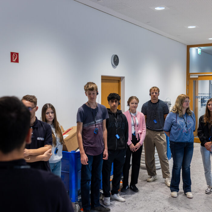 Eine Gruppe von Auszubildenden steht in der Additive Fertigung. Ein Ausbilder erzählt den neuen Auszubildenden etwas über die Abteilungen.