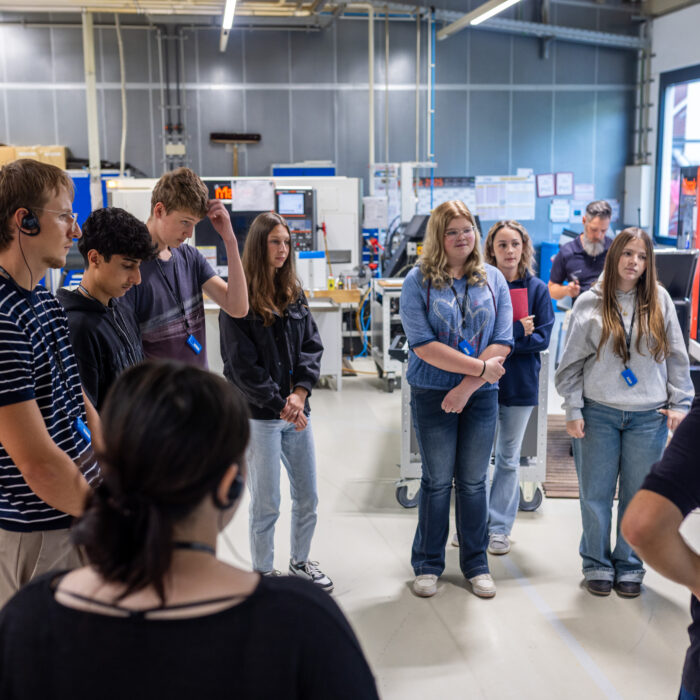 Eine Gruppe von Auszubildenden befinden sich in der Produktion. Ein Auszubildender erklärt was in der Abteilung Dreh und Frästeile bearbeitet wird.
