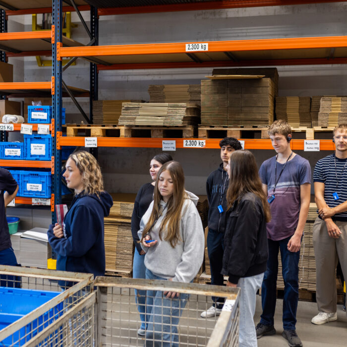 In der Produktionsabteilung ist eine Gruppe von Auszubildenden zu sehen. Einer der Auszubildenden erklärt den anderen die Arbeitsprozesse.