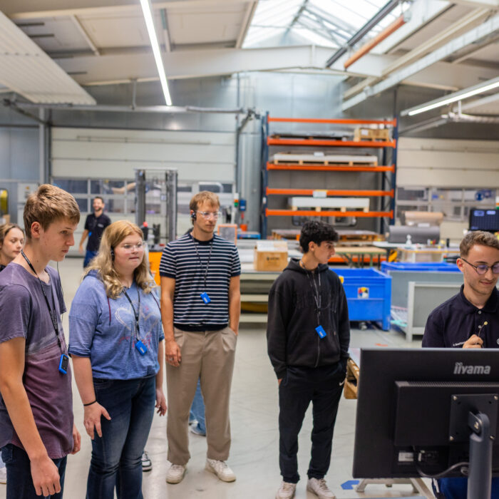 In der Produktionsabteilung befindet sich eine Gruppe von Auszubildenden. Ein Auszubildender erklärt den anderen das Programm einer Maschine und zeigt dessen Anwendung im Arbeitsprozess.