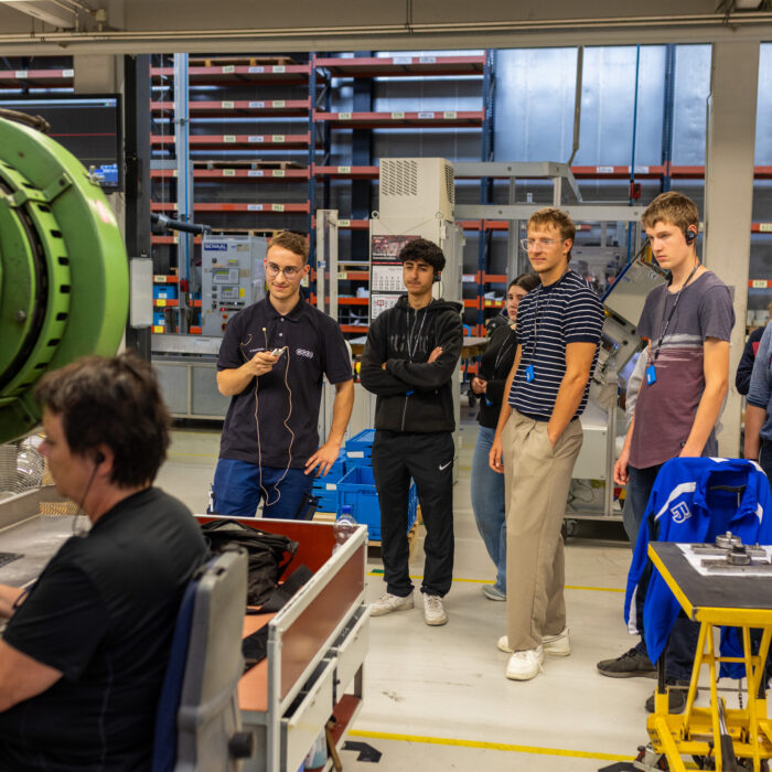 In der Produktion befindet sich eine Gruppe von Auszubildenden. Es wird eine Stanzmaschine in Betrieb betrachtet.