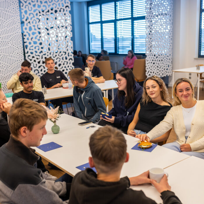 Auszubildende sitzen an einem Tisch in der Kantine um zu Frühstücken. Viele Auszubildende unterhalten sich oder lächeln in die Kamera.