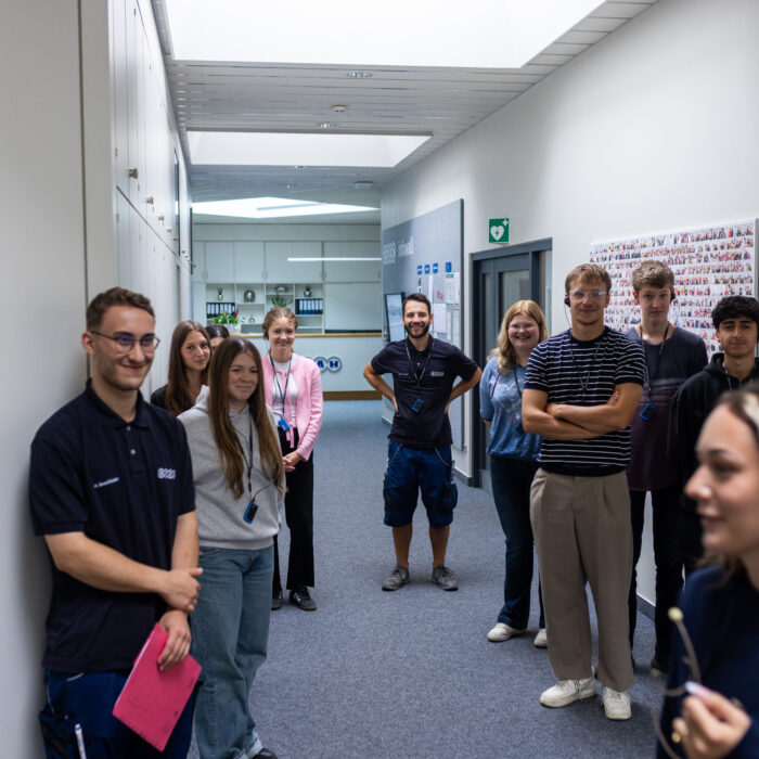 Eine Gruppe von Auszubildenden steht in einem Gang in der Verwaltung. Eine Auszubildende gibt den neuen Auszubildenden eine Führung durch das Unternehmen und erzählt ihnen etwas.