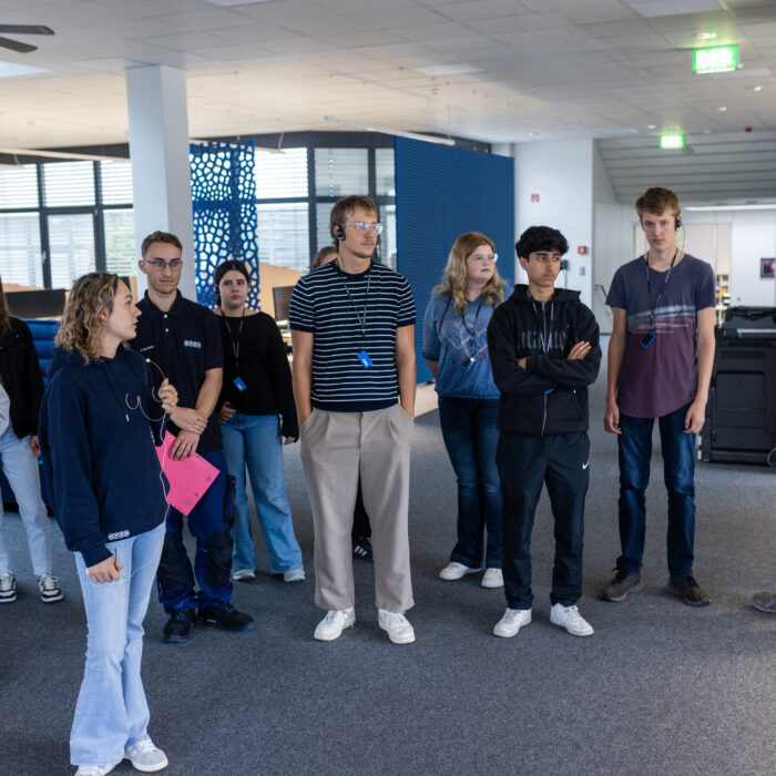 Eine Gruppe von Auszubildenden steht in der Verwaltung. Eine Auszubildende erzählt den neuen Auszubildenden bei einer Führung etwas über die Abteilungen.