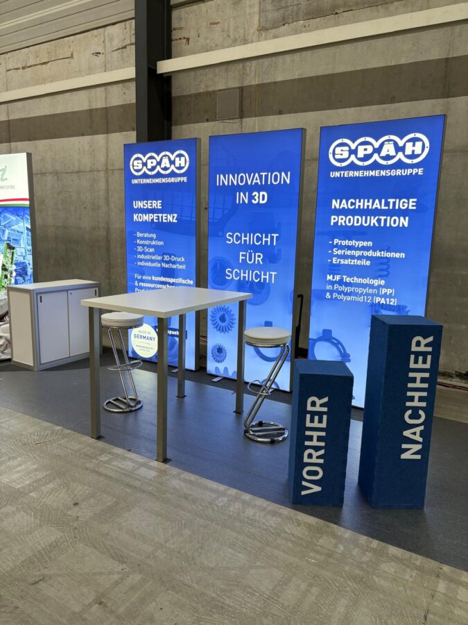 Späh Messestand mit einem Tisch und zwei Stühlen. Hinten sind drei Wände mit den Titeln "Unsere Kompetenz", "Innovation in 3D" und "nachhaltige Produktion".