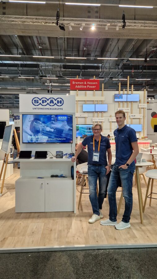 Zwei lächelnde Späh-Mitarbeiter sind auf einem Messestand. Auf dem Messestand ist ein Bildschirm zu sehen in dem es um die Fertigungsart Drehen und Fräsen geht.