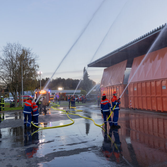 Mehrere Gruppen von Mitarbeitern der Jugendfeuerwehr geben einen Strahl ab mit dem Wasserschlauch auf die Überdachung der Müllcontainer.