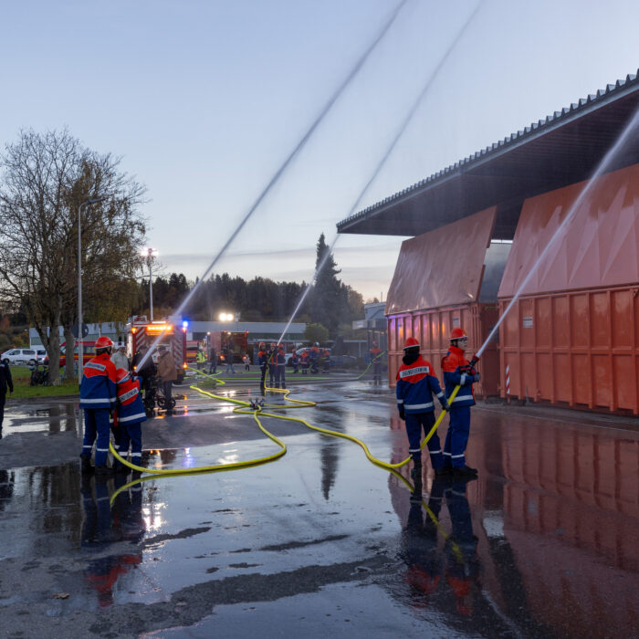 Mehrere Gruppen von Mitarbeitern der Jugendfeuerwehr geben einen Strahl ab mit dem Wasserschlauch auf die Überdachung der Müllcontainer.
