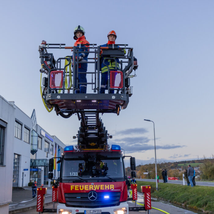 Zwei Mitarbeiter der Jugendfeuerwehr stehen auf dem Korb der Drehleiter. Das Feuerwehrfahrzeug steht vor dem Warenausgang.