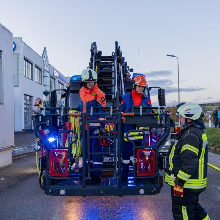 Der Korb der Drehleiter ist ganz tief eingestellt. Zwei Mitarbeiter der Jugendfeuerwehr stehen in dem Korb und ein Mitarbeiter der Feuerwehr steht daneben. Das Feuerwehrfahrzeug steht vor dem Warenausgang.