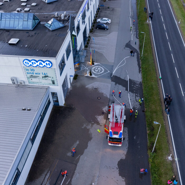 Ein Feuerwehrfahrzeug steht vor dem Haupteingang vom Späh. Um das Fahrzeug herum sind Mitarbeiter der Jugendfeuerwehr die einen Wasserschlauch halten. An der Seite stehen Zuschauer.