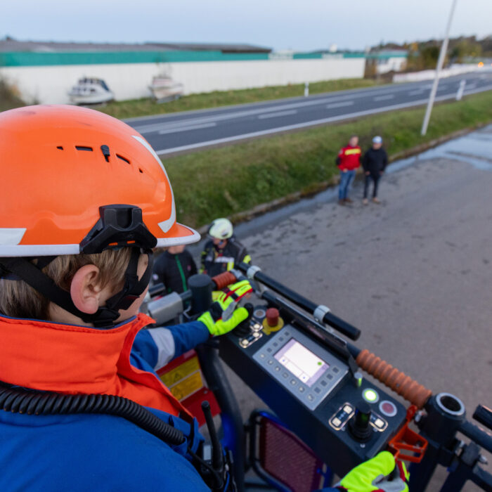Ein Junge aus der Jugendfeuerwehr mit orangenem Helm bedient eine Maschine. An der Seite steht ein Mann aus der Feuerwehr und zwei Zuschauer.