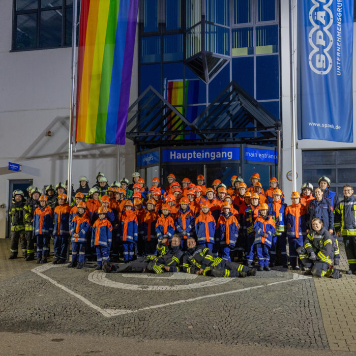 Gruppenfoto zwischen der Jugendfeuerwehr und Feuerwehr vor dem Haupteingang von Späh. Die Jugendfeuerwehr und Feuerwehr sind jeweils einheitlich in ihrer Bekleidung.