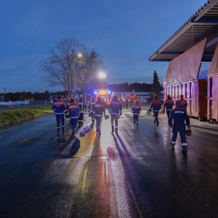 Mehrere Kinder aus der Jugendfeuerwehr und Erwachsene aus der Feuerwehr sind auf dem Späh Gelände für eine Übung. Sie gehen zusammen in die Richtung des blinkenden Feuerwehrfahrzeugs.
