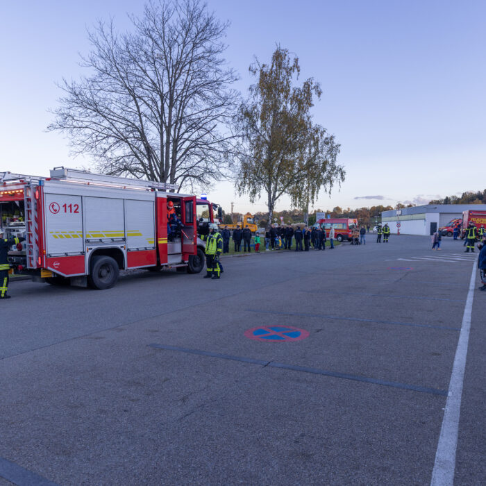 Ein Feuerwehrfahrzeug ist auf dem Späh Gelände. Davor stehen drei Menschen aus der Feuerwehr. Im Hintergrund sind Zuschauer die in Richtung des Fahrzeugs schauen.