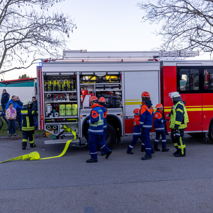 Mehrere Kinder aus der Jugendfeuerwehr und 2 Männer aus der Feuerwehr stehen vor dem offenen Feuerwehrfahrzeug. Ein Mann erklärt den 2 Jungen etwas und die anderen bereiten etwas vor.