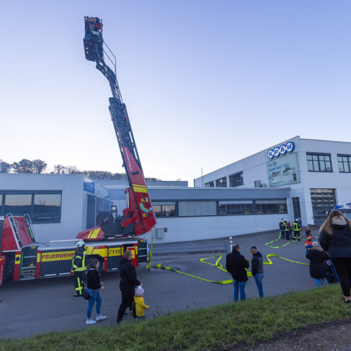 Vor dem Späh Gebäude stehen zwei Feuerwehrfahrzeuge. Die Schläuche zum Löschen sind ausgerollt und die Drehleiter bei dem linken Fahrzeug ist weit in der Luft. Zurückgezogen schauen mehrere Menschen dabei zu.