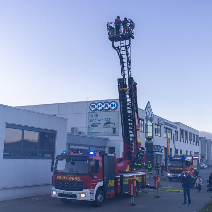 Vor dem Warenausgang steht ein Feuerwehrfahrzeug. Ein Mann aus der Feuerwehr fährt die Drehleiter nach oben. Im Korb der Drehleiter sind zwei Kinder aus der Jugendfeuerwehr. Mehrere Menschen schauen dabei zu.