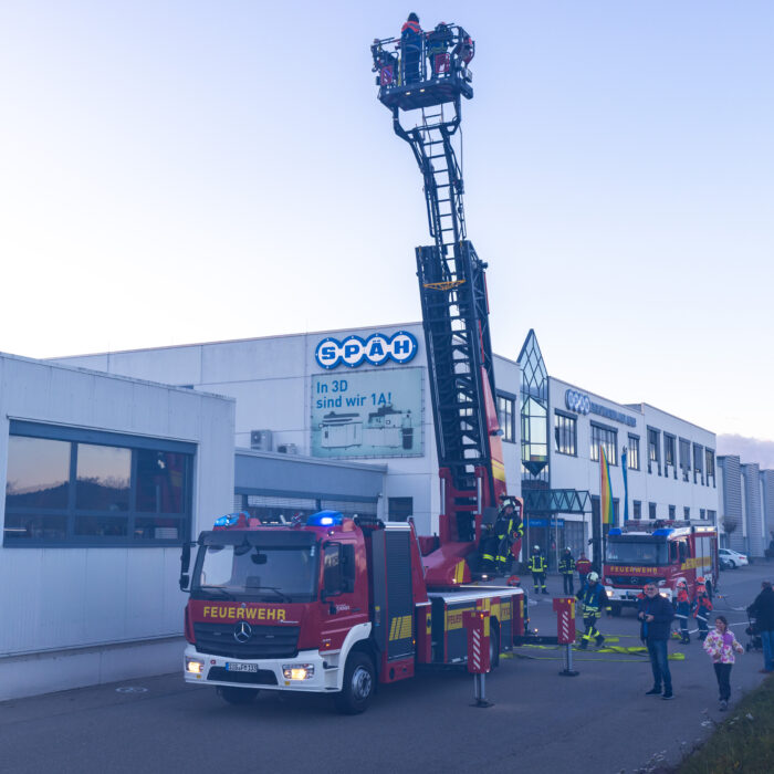 Vor dem Warenausgang steht ein Feuerwehrfahrzeug. Ein Mann aus der Feuerwehr fährt die Drehleiter nach oben. Im Korb der Drehleiter sind zwei Kinder aus der Jugendfeuerwehr. Mehrere Menschen schauen dabei zu.